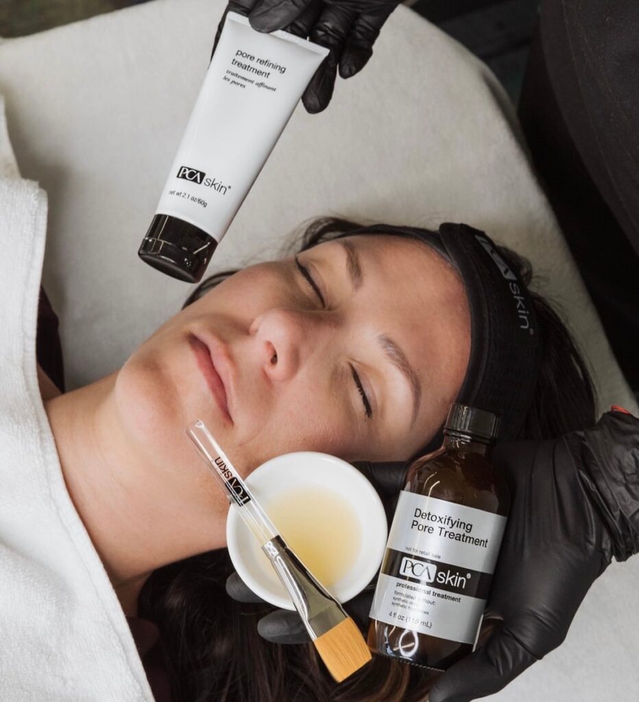 An image showing a hand wearing a black glove holding a tube of PCA Skin professional skin care treatment, which appears to be a detoxifying pore treatment, placed next to a woman's face as she receives a skincare treatment.