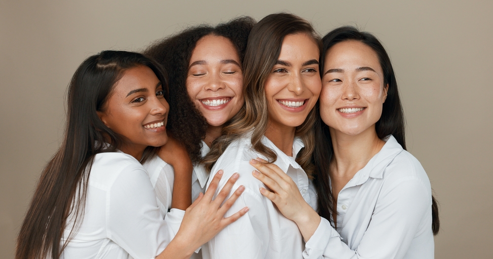 Four smiling women of diverse ethnicities wearing white shirts posing together against neutral background