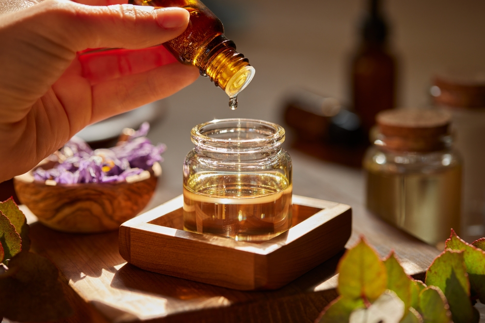 Hand applying golden essential oil or hair serum from dropper bottle into glass jar surrounded by natural ingredients
